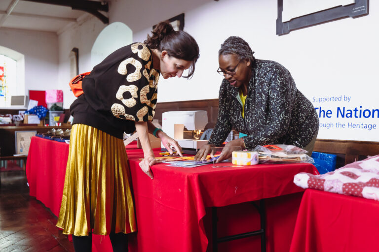 Two women leaning over a table covered in a red cloth, setting out materials for a Fun Palace activity.