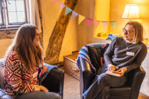 Two women are seated in a cozy, warmly lit room having a friendly conversation. One woman with long brown hair, wearing a red and white patterned sweater and a name tag that reads "Annie," is smiling and engaged in the conversation. The other woman, wearing glasses, a gray sweater, and a name tag that reads "Hanya," is holding a notebook and pen. Behind them is a window, colorful bunting, a wicker basket, and a lamp, contributing to the relaxed and inviting atmosphere.