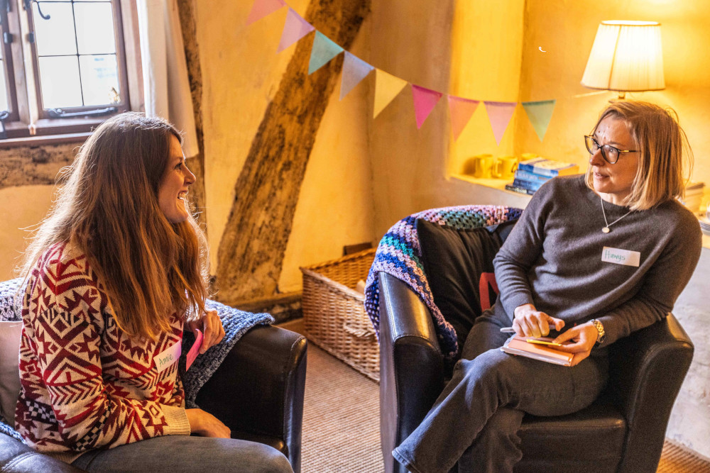 Two women are seated in a cozy, warmly lit room having a friendly conversation. One woman with long brown hair, wearing a red and white patterned sweater and a name tag that reads "Annie," is smiling and engaged in the conversation. The other woman, wearing glasses, a gray sweater, and a name tag that reads "Hanya," is holding a notebook and pen. Behind them is a window, colorful bunting, a wicker basket, and a lamp, contributing to the relaxed and inviting atmosphere.