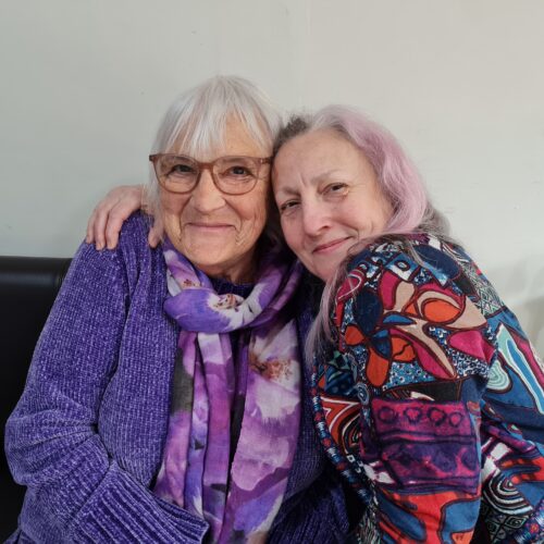 Two white women hugging, one with white hair and glasses, one with pink and white hair.