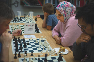 Someone wearing a hijab and pink jumper concentrates on a chess game in a library.