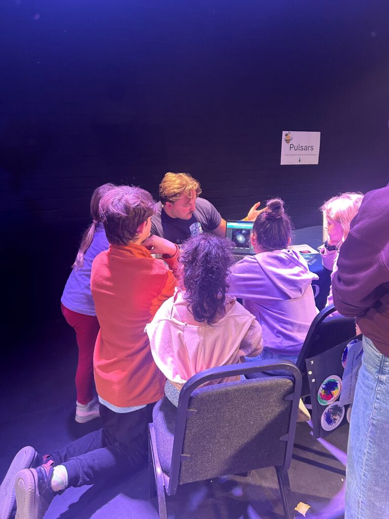 A group of children gather around a table where an adult is animatedly explaining something, pointing towards a laptop screen displaying an image of space. The scene is warmly lit with purple and blue lighting. A sign on the dark wall behind them reads “Pulsars.” The atmosphere appears lively and engaging, likely part of a workshop or educational event.