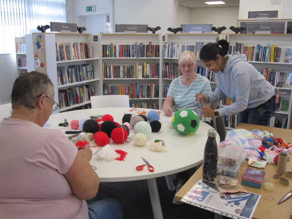 Picture of Cristina demonstrating a craft with wool, with two attendees sat at the table, which has balls of wool in the middle.