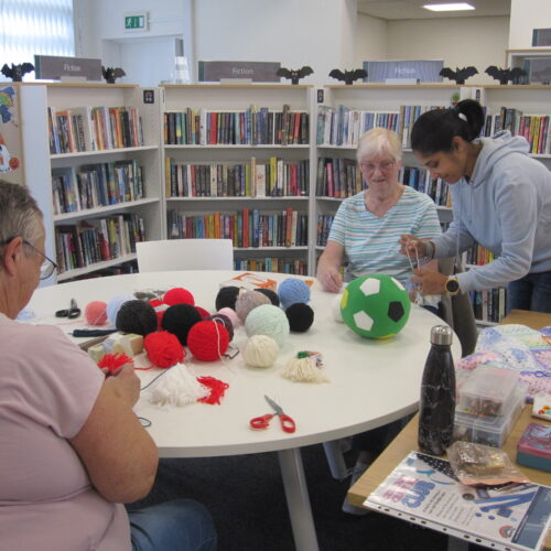 Picture of Cristina demonstrating a craft with wool, with two attendees sat at the table, which has balls of wool in the middle.