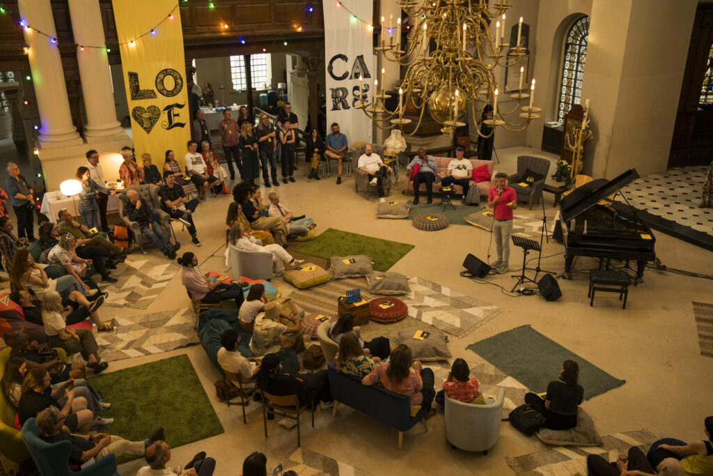 Photo of people gathering in a large room, wither sitting on sofas or standing. There's a person at the microphone. In the room is a chandelier, fairy lights, and love and care are written on the walls.