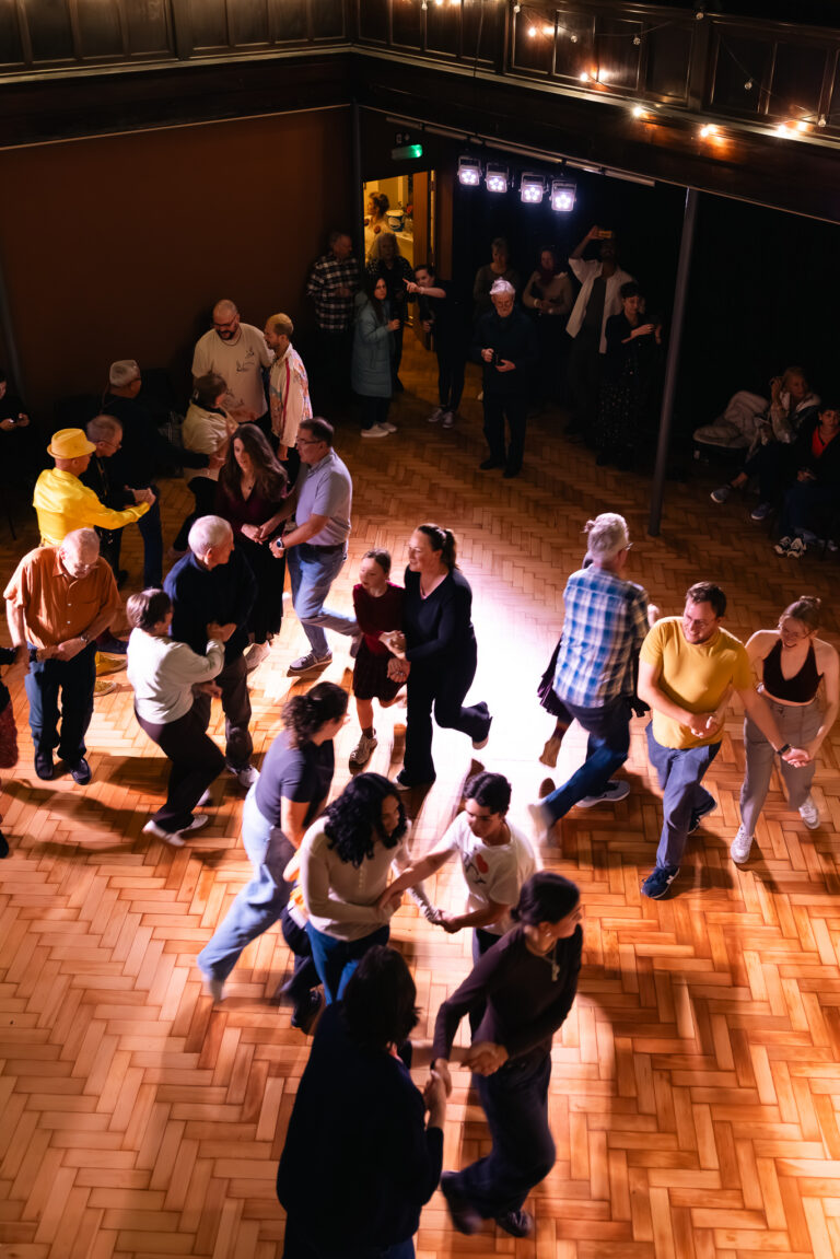 Seen from above, pairs of people dance together across a wooden floor in a hall strung with lights.