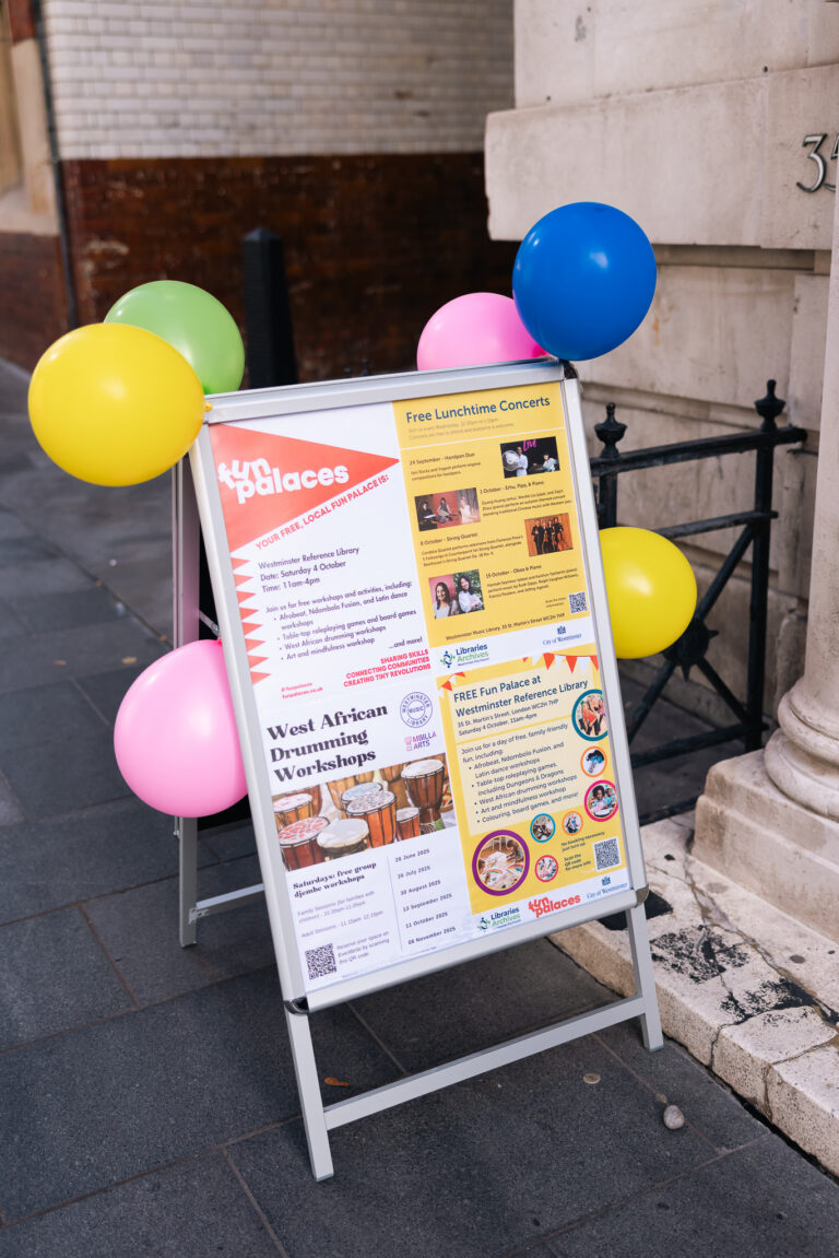 A Fun Palaces A-board poster outside a building, decorated with bright yellow, pink, green and blue balloons.