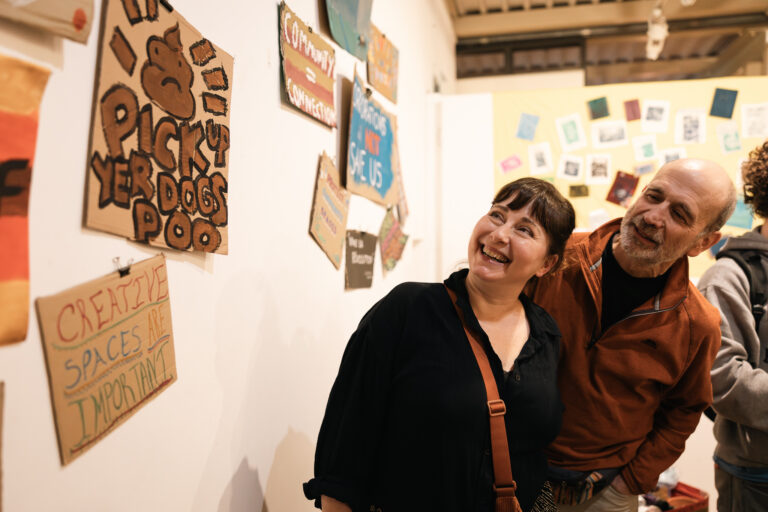 Two people laughing together in front of a wall of handmade cardboard placards at a Fun Palaces event.