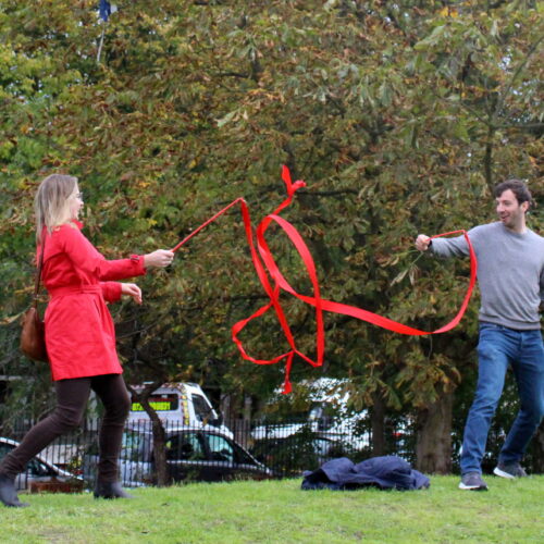 picture of a women in a red jacket and a man in a grey sweatshirt and jeans, having fun using rhythmic gymnastics ribbons